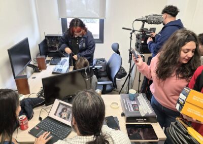 Seis personas, entre ellas estudiantes, ayudantes y profesores, están en una sala pequeña con computadores, cámaras y cables. Algunos trabajan en un PC, mientras otros revisan equipos de audio y video en un ambiente desordenado de preparación técnica.