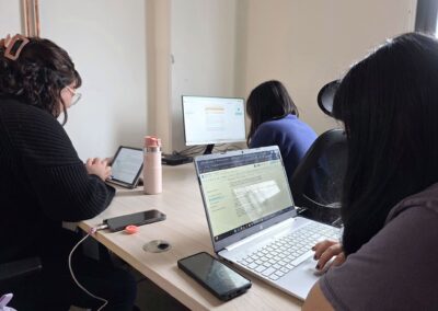 Tres estudiantes de periodismo escriben sobre una mesa en forma de “ele” en una oficina con fondo blanco del Instituto de Comunicación de la UACh. Una ocupa un IPad, otra un computador portátil y otra con un computador de escritorio.
