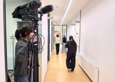 Una estudiante de periodismo sostiene un trípode con micrófonos y cables en el umbral de una puerta mirando hacia el pasillo, mientras otras tres caminan hacia el fondo a la sala del evento.