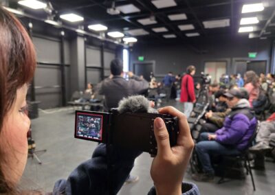 Primer plano de una estudiante de periodismo, con cámara en mano, comienza a fotografiar al público en la actividad. Sosteniendo. Hay un grupo de asistentes sentados en un auditorio, con varias cámaras instaladas alrededor.