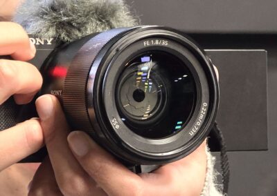 Primer plano de una cámara Sony en manos de una estudiante de periodismo. Se distingue la lente de 35 mm con apertura f/1.8 y reflejos de luces en el cristal.