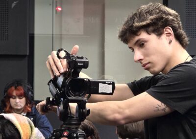 Un estudiante de periodismo ocupa una cámara Sony montada sobre un trípode. Está ajustando el equipo con atención mientras al fondo a otra persona monitoreando el audio.