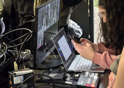 Varias personas trabajan en un puesto de control con una pantalla, un computador portátil, un mezclador de video y varios cables conectados. Una persona revisa un celular mientras monitorea la transmisión.