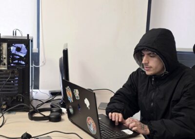 Un estudiante de periodismo, vestido de negro, del equipo de medios sociales está editando frente a un computador portátil, sobre una mesa en una habitación blanca con ventanas grandes detrás de él.